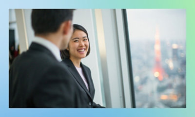 ビジネス服を着た 2 人が、背景にぼやけた塔を含む都市の景観が見える窓の近くに立っています。