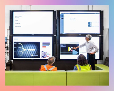 A man giving a presentation to a group of people with large screens in the background.
