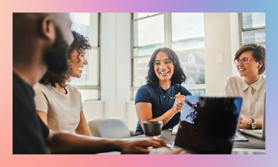 Three individuals engaged in a meeting around a table with laptops and coffee cups, in a room with large windows.