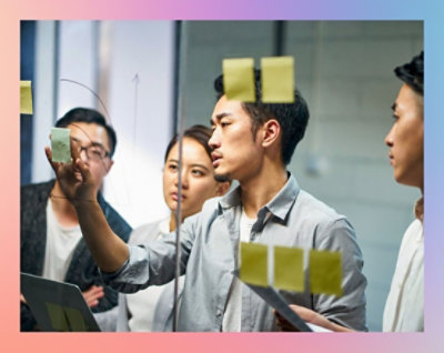 Čtyři osoby spolupracují v kanceláři. Muž ukazuje na samolepicí papírky na skleněné tabuli, zatímco ostatní jej sledují.