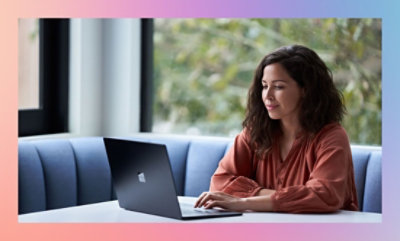 Person in a terracotta blouse working on a laptop at a white table with a blue couch in the background.