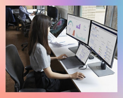 Žena sedí u stolu v kanceláři a používá notebook připojený ke dvěma monitorům, které zobrazují různé typy grafů s daty.