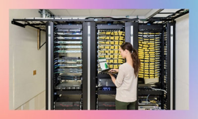 A woman using a laptop while standing in front of an open server rack in a data center.