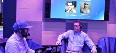 Two headset-wearing individuals sit and talk during a podcast recording, with a screen behind showing speaker profiles.