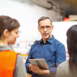A man holds a tablet and talks to four people, two of whom are wearing orange safety vests, in an indoor setting.