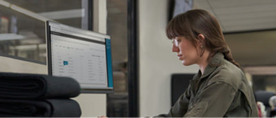 A woman with brown hair and glasses, wearing a green shirt, looks at a computer screen.