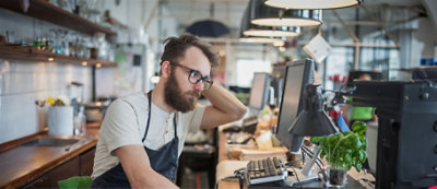 גבר מזוקן מרכיב משקפיים ולובש סינר כחול, יושב ליד שולחן מבולגן בסביבת עבודה מוארת היטב