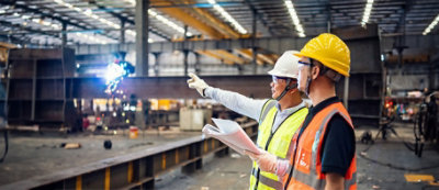 Two workers wearing hard hats and safety vests stand in a large industrial warehouse