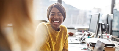 Person in a yellow sweater sitting at a desk with multiple computer screens in a bright office setting.