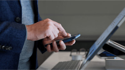 Hands holding a mobile phone in front of a desktop monitor.