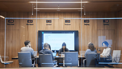A group of people meeting in a conference room with a world clock on the wall.