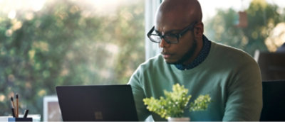 眼鏡をかけた男性がテーブルに座ってノート PC で作業しており、近くには植物とペンが置いてあります。