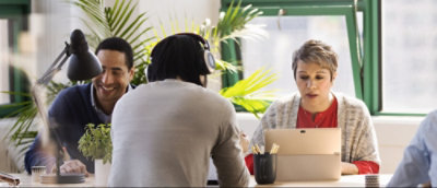 Three individuals working at a desk with laptops and headphones in an office environment with plants in the background.