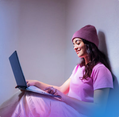 Woman in a knit hat uses a laptop while sitting against a wall
