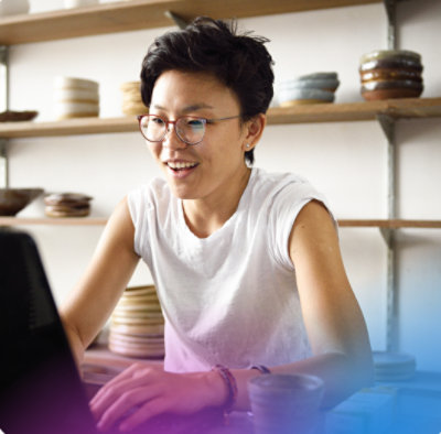 Smiling person with glasses works on a laptop in a pottery studio