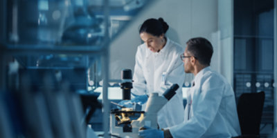 Two scientists in white lab coats work together in a laboratory, examining samples under a microscope