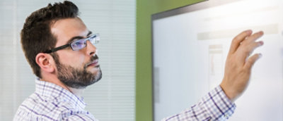 Person in a plaid shirt gesturing towards a whiteboard in a bright room.