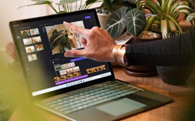 A person uses a touchscreen laptop to edit photos, with several plants in the background.