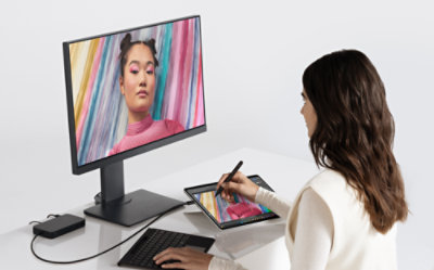 A person works on a tablet with a stylus while looking at a colorful portrait displayed on a connected computer monitor.