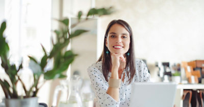 Žena sedi za stolom na kojem se nalazi laptop, u blizini je biljka, i zamišljeno gleda u daljinu.