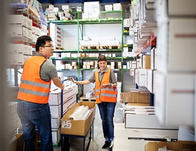Two warehouse workers in orange safety vests organize boxes and check inventory on a clipboard 