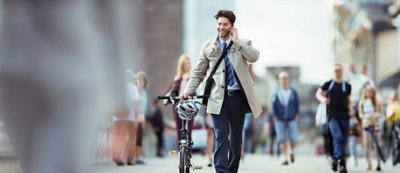 Mężczyzna w garniturze i trenczu jadący na rowerze ulicą miasta i rozmawiający przez telefon komórkowy.