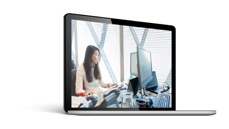 デスクで PC を使用して仕事をする人物が画面に表示されたノート PC
