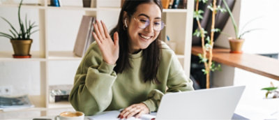 Une personne en sweat vert fait un signe de la main à l’écran d’un ordinateur portable, assise à un bureau devant des étagères avec des plantes et des livres.