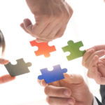 Four people each hold a different colored puzzle piece, working together to connect them.