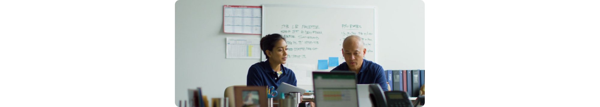 Two people sitting at a desk in front of a whiteboard.