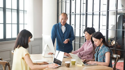 Četiri osobe u dobro osvetljenoj kancelariji rade zajedno za stolom na kojem se nalaze laptop računari i jedan stoni računar.