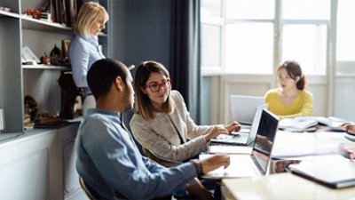 Four people are working together at a table with laptops and a desktop computer in a well-lit office space.