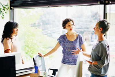 Tri žene u razgovoru u zatvorenom kancelarijskom prostoru, pri čemu jedna drži beležnicu a druge dve stoje i slušaju.