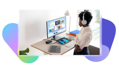 Eine Person, die an einem Schreibtisch steht, ohrumschließende Kopfhörer trägt und auf einen Desktopmonitor schaut