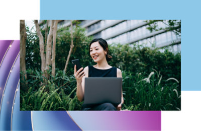 Nasmejana žena sa sedom kovrdžavom kosom i naočarima koja gleda u pametni telefon i stoji na gradskom trotoaru.