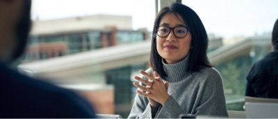 A woman in a grey turtleneck and glasses with her hands together.