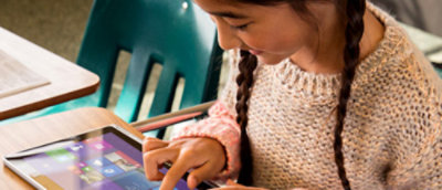 A girl with braided hair uses a tablet at a school desk, focused on the screen.