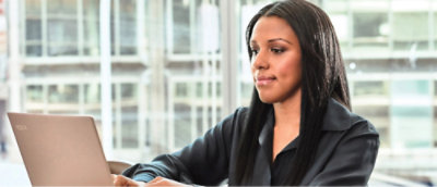 Une femme assise à un bureau, concentrée sur son ordinateur portable, entourée d'un espace de travail bien rangé.