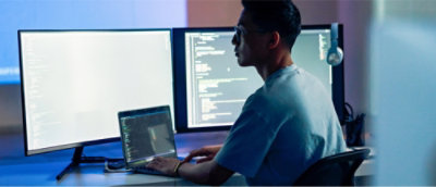 薄暗い部屋で、2 つの大きなモニターにコードを表示してノート PC で作業している人。