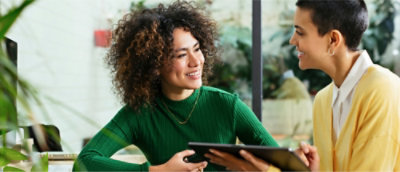 Deux personnes assises à une table discutent en souriant. L’une d’elles tient une tablette. Environnement intérieur avec des plantes en arrière-plan.