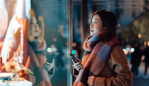Una persona con una bufanda grande sostiene un teléfono móvil y mira el escaparate de una tienda iluminado por las luces de la ciudad.