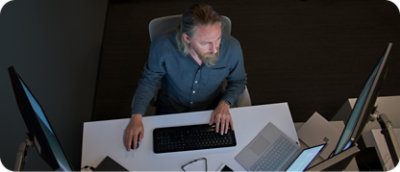 Muž s bradou sedí za stolom a používa prenosný a stolový počítač s viacerými monitormi.