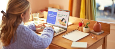 En kvinna i blå tröja använder en bärbar dator på ett skrivbord i trä, med en öppen anteckningsbok, en penna och en växt