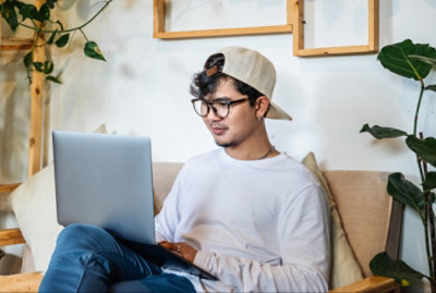Persona con gafas y gorra sentada en un sofá usando un portátil, rodeada de plantas.