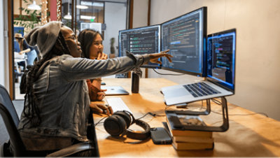 Two people working together at a desk with two monitors and a laptop.