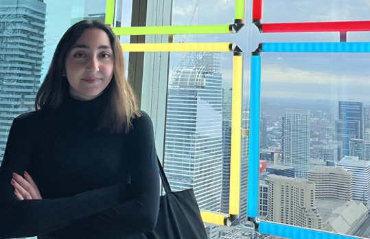Celina Tuffaha stands with her arms crossed in front of a large window showing a panoramic cityscape