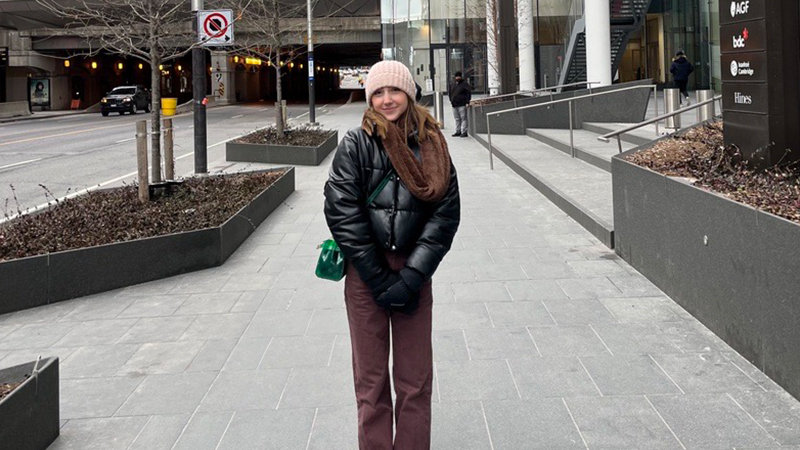 Celina Tuffaha, vêtue d’une veste noire et d’un pantalon marron, se tient sur un trottoir de la ville, un sac vert à la main, avec des panneaux de signalisation et des bâtiments urbains en toile de fond