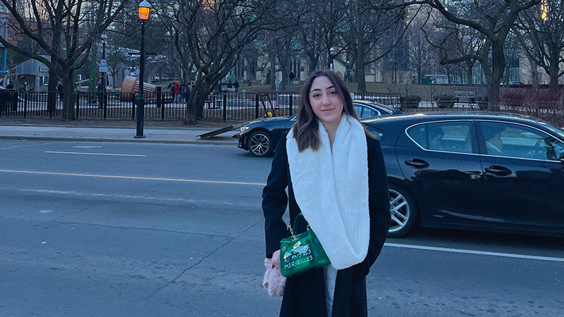 Celina Tuffaha in a dark coat and white scarf stands on a sidewalk holding a green ‘MARC JACOBS’ bag