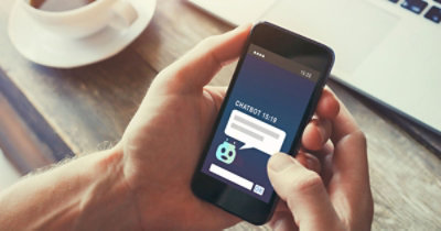 Osoba drži pametni telefon na kojem se vidi razgovor sa botom za ćaskanje i poruka u 15:19. U pozadini se vidi laptop.