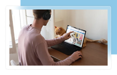 Person wearing headphones uses a laptop at a desk, pointing at the screen while a cat sits beside the laptop.